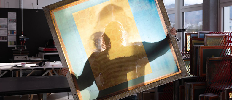 Eine Person hält in einem hellen Atelier einen grossen, rechteckigen, durchscheinenden Rahmen mit farbigen Schichten vor sich, sodass ihr Schatten auf dem Material sichtbar wird. Im Hintergrund sind Fenster, Arbeitstische und gestapelte Siebdruckrahmen zu sehen