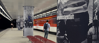 Schwarz-weiss gestaltete Ausstellungsgrafiken auf Säulen in einem Bahnhof der Zentralbahn, gestaltet von Nodo Creative Hub; im Hintergrund fährt ein roter Zug ein, während Passant:innen auf dem Bahnsteig stehen.
