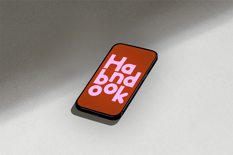 Ein Handy mit weinrotem Hintergrund, auf dem in pinker Schrift Handbook steht. Das Handy liegt auf einer grauen Fläche, die zur hälfte mit Schatten bedeckt ist. 
