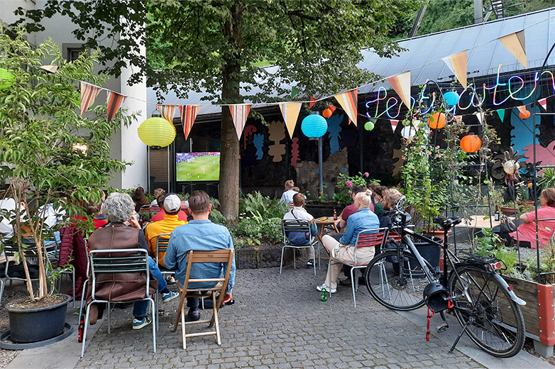 Bunt gestaltete Plakate und Drucksachen im öffentlichen Raum sowie Szenen eines lebendigen Quartiertreffs mit Menschen, Pflanzen und gemeinschaftlichen Aktivitäten im Aussenbereich.