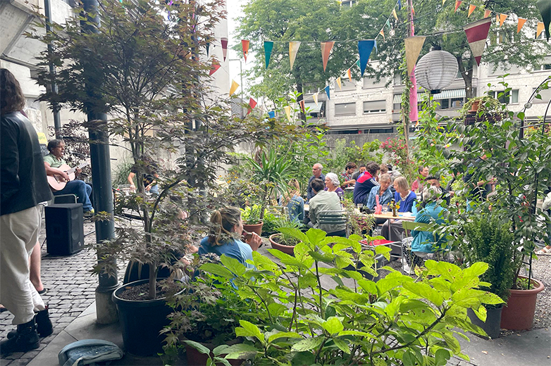 Bunt gestaltete Plakate und Drucksachen im öffentlichen Raum sowie Szenen eines lebendigen Quartiertreffs mit Menschen, Pflanzen und gemeinschaftlichen Aktivitäten im Aussenbereich.