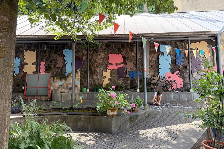 Bunt gestaltete Plakate und Drucksachen im öffentlichen Raum sowie Szenen eines lebendigen Quartiertreffs mit Menschen, Pflanzen und gemeinschaftlichen Aktivitäten im Aussenbereich.