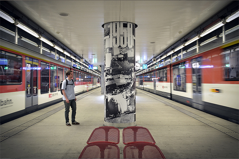 Bahnsteig in der Zentralbahnstation Allmend/Messe, im Zentrum eine Betonsäule mit der Aufschrift „1950“ und historischen Schwarzweissbildern; ein Zug fährt ein, eine Person steht neben der Säule.