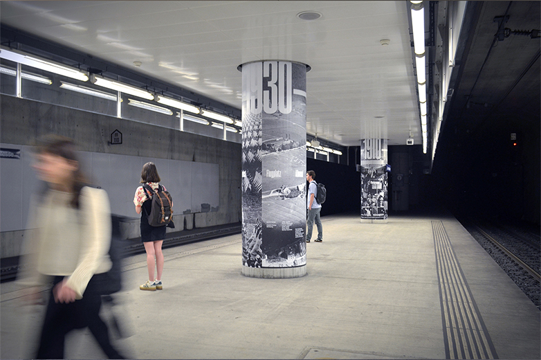 Mehrere Personen bewegen sich durch die Unterführung der Zentralbahnstation, typografisch gestaltete Säulen mit Jahreszahlen wie „1930“ zeigen historische Bildmotive, unter anderem zum Flugplatz Allmend.