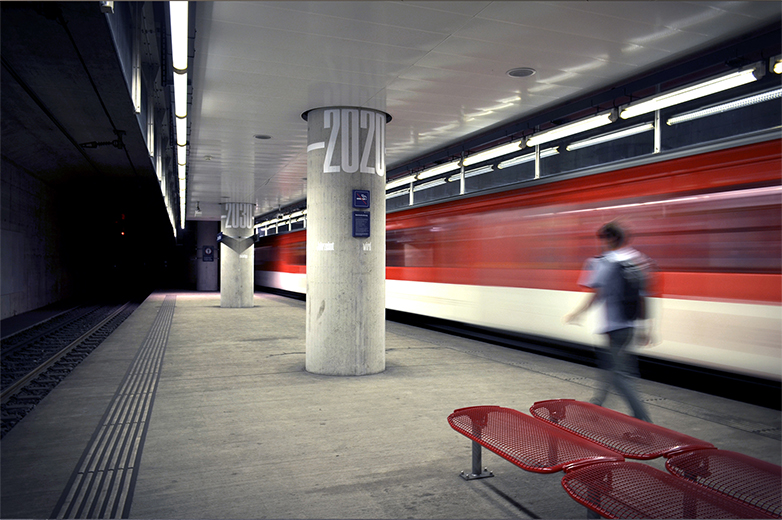Zentralbahn-Station mit durchfahrendem Zug, im Fokus eine Betonsäule mit der typografischen Jahreszahl „–2020“; eine Person läuft am Bahnsteig entlang.