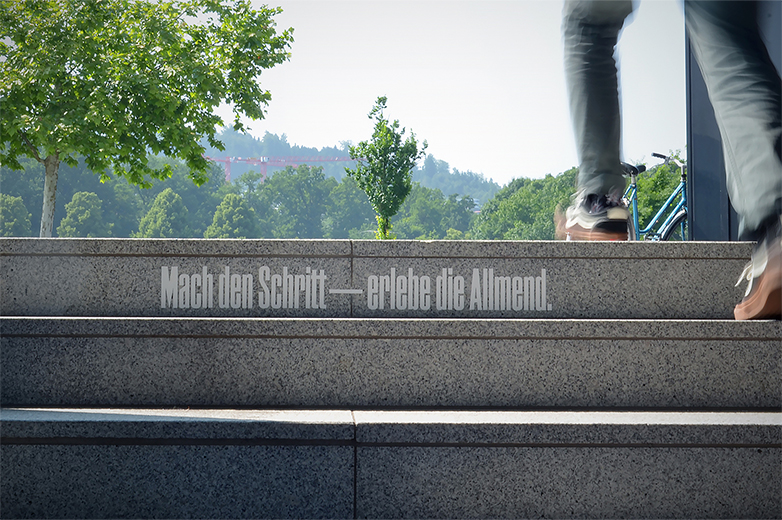 Zwei Personen steigen eine Freitreppe im Aussenbereich hinauf, auf einer Stufe steht typografisch „Mach den Schritt — erlebe die Allmend“, im Hintergrund Bäume und Fahrräder.