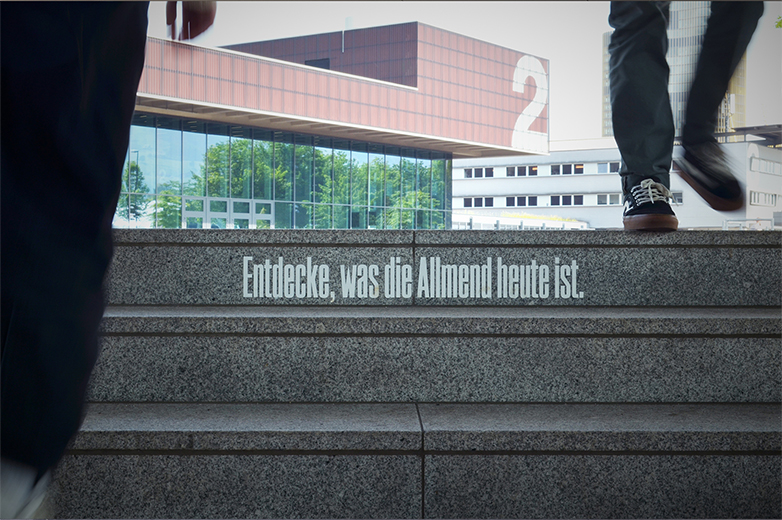 Zwei Personen steigen eine Aussen-Treppe vor einem modernen Gebäudekomplex hoch, auf der Stufe ist zu lesen: „Entdecke, was die Allmend heute ist“.