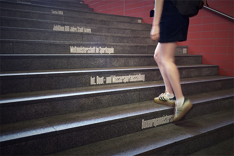Gestalterische Interventionen im öffentlichen Raum rund um die Allmend/Messe Luzern, mit Beschriftungen, historischen Bezügen und Menschen in Bewegung an Haltestellen, Treppen und Zugängen.