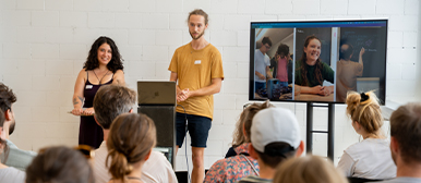     Präsentation vor Publikum: Zwei Personen präsentieren vor einem sitzenden Publikum in einem hellen Raum. Hinter ihnen zeigt ein Bildschirm Fotos ihres Projekts.