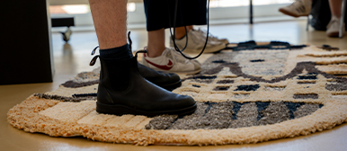     Teppich: Nahaufnahme der Beine einer Person, die auf einem runden, handgefertigt wirkenden Teppich mit abstraktem Muster in Beige, Grau und Dunkelblau steht.