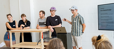     Gruppenpräsentation: Fünf Personen stehen hinter einer Holzkonstruktion und präsentieren gemeinsam vor einem Publikum. Rechts zeigt ein Bildschirm Informationen zum Thema Eco-Social Design.