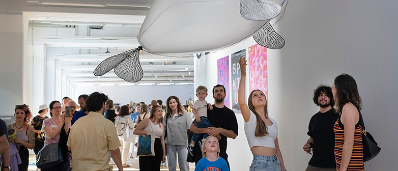 Menschengruppe betrachtet eine fliegende Skulptur an der Werkschau.