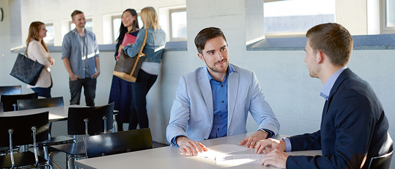 Zwei Männer haben eine Besprechung und im Hintergrund ist eine Gruppe Studierender zu sehen
