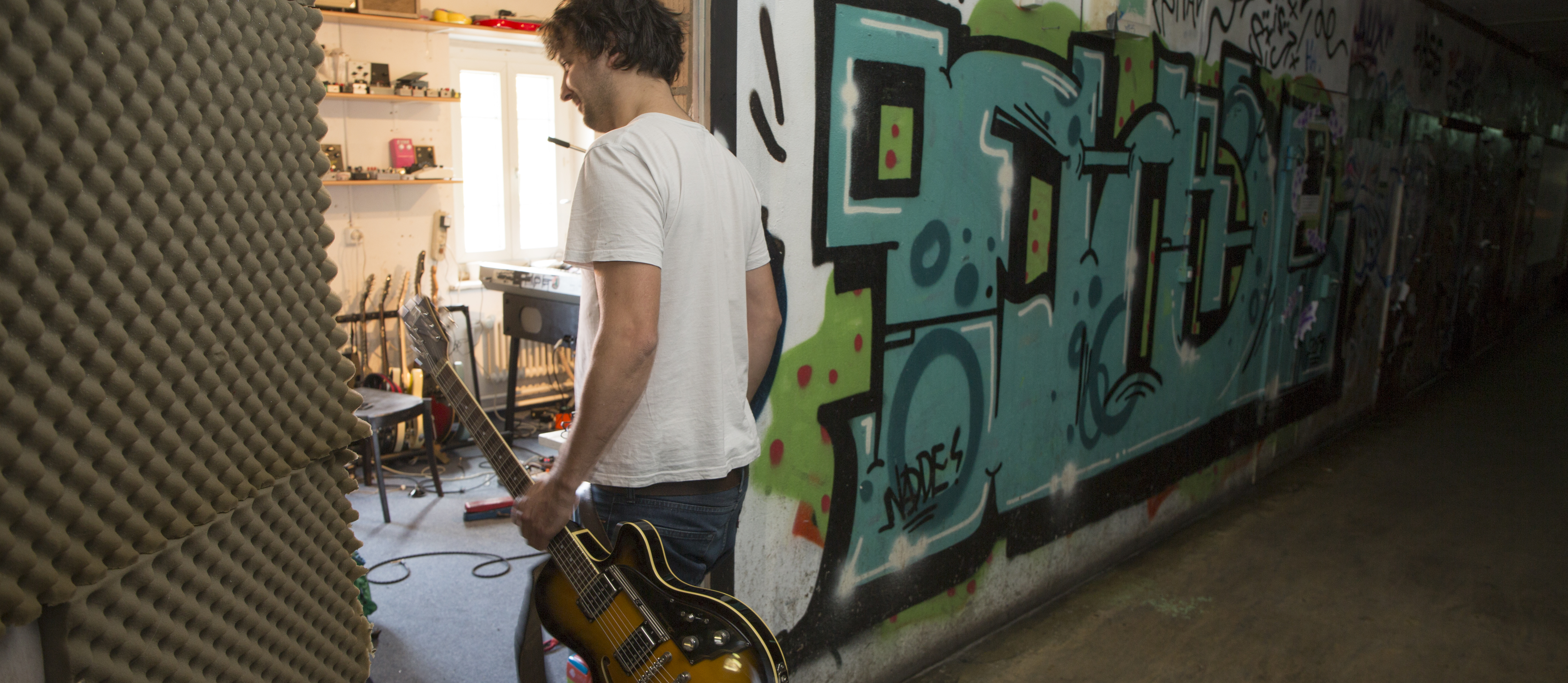 Ein junger Mann mit E-Gitarre tritt in ein Proberaum ein. 
