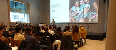 Eine Gruppe interessierter jungen Personen folgt der Präsentation eines Mannes auf der Bühne