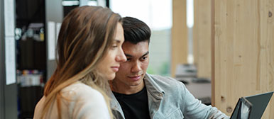 Ein Mann und eine Frau sitzen vor einem grossen Fenster und schauen gemeinsam auf einen Laptop.