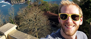 Ein Selfie eines jungen Mannes mit Sonnenbrille und im Hintergrund sind Bäume und ein knallblaues Gewässer zu sehen