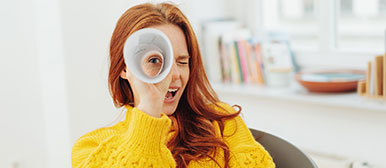 Eine junge Frau mit roten Haaren und einem gelben Pullover hält ein aufgerolltes Blatt Papier vor ihr Auge und schaut hindurch wie durch ein Fernrohr