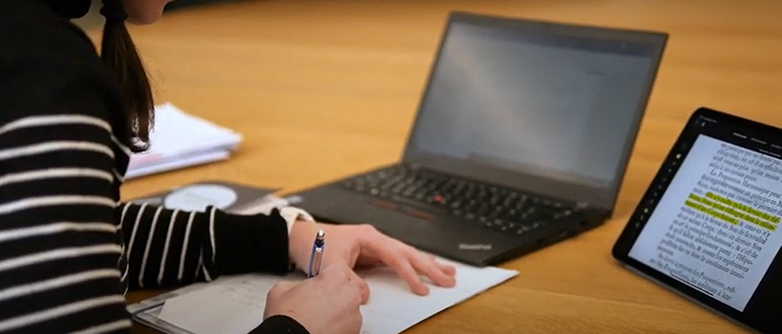 Screenshot eines Videos: Hände schreiben etwas auf einem Papier, auf dem Pult stehen noch einen Laptop und ein Tablet. 