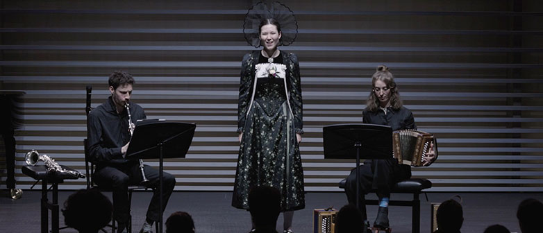 Drei Musiker:innen auf der Bühne: Klarinettist, Sängerin in traditioneller Tracht und Akkordeonspielerin während eines Konzerts.