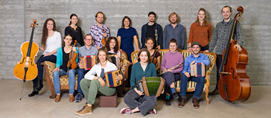 Ein Portrait mit den acht Mitglieder des Volksmusik-Ensembles Alpinis. Die Mitglieder stehen draussen vor einem Baum und haben ihre Instrumente in der Hand. 