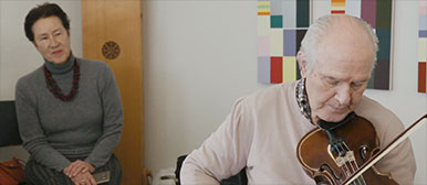 Ein demenzkranker Mann spielt Violine, im Hintergrund sitzt eine Frau. 