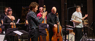Mehrere Musiker:innen stehen auf einer Bühne, Cello, Posauen und Geige sind sichtbar