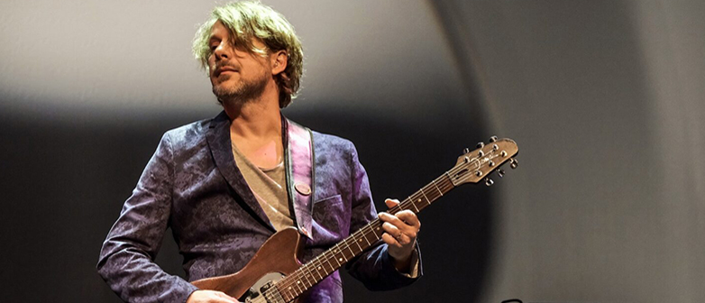 Kalle Kalima steht mit einer E-Gitarre auf der Bühne. ER trägt einen violetten Anzug
