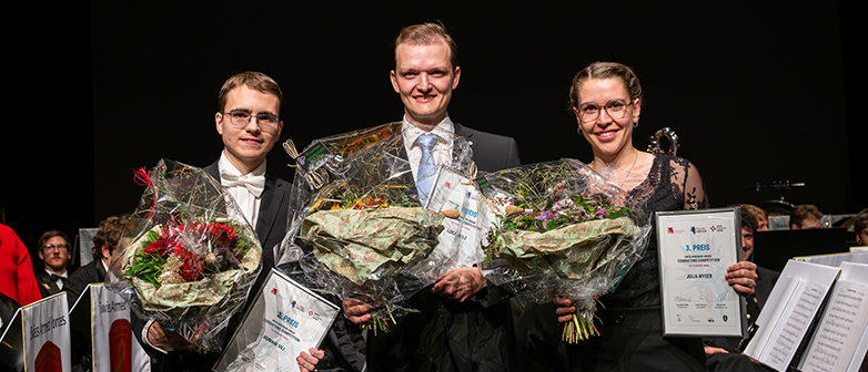 Drei Preisträger:innen der Conducting Competition 2026 stehen mit Blumensträussen und Urkunden auf der Bühne.