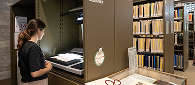 Eine Frau steht in der Musikbibliothek am Scanner, vor ihr sind mehrere Regal mit Büchern