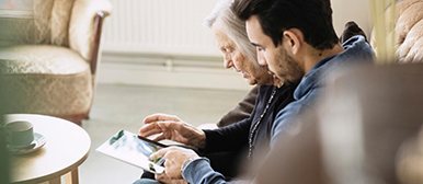 Ein junger Mann und eine ältere Frau sitzen nebeneinander auf einem Sofa und betrachten das Display eines Tablets