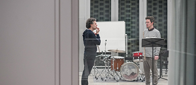 Durch ein Fenster sind zwei Musiker in einem Überaum sichtbar im Gespräch