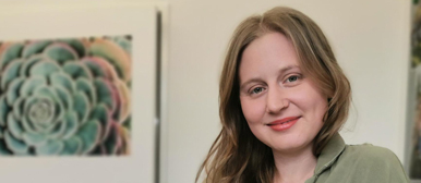 Eine Frau die lächelnd in die Kamera schaut und im Hintergrund ist ein Bild einer Pfanze zu sehen