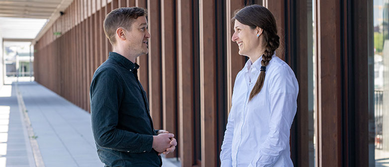 Zwei Personen stehen auf einer Terrasse neben einem Gebäude mit Glasfront und sprechen miteinander. 