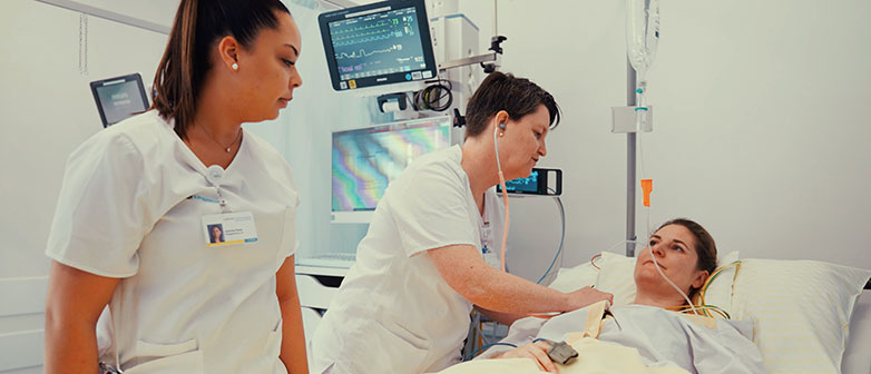 Pflegefachpersonen untersuchen eine Patientin im Spitalbett, umgeben von medizinischen Geräten.