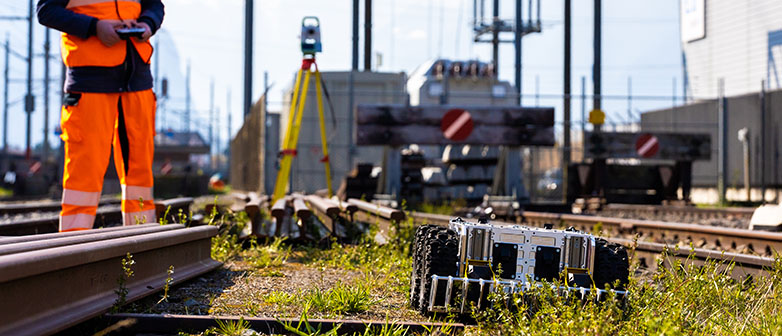 Autonome Systeme und Robotik: Mann in Arbeitskleidung steht auf Bahngleis mit dem Controller für den mobilen Roboter nebendran in der Hand