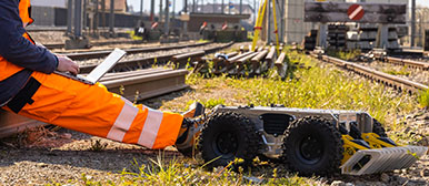 Mobile Robotik: Mann in Arbeitskleidung sitzt mit Laptop auf Bahngleis und kleiner Roboter sitzt daneben