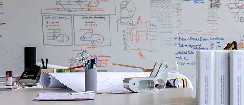 Produktentwicklung: Whiteboard mit Berechnungen und Skizzen, Pult mit Stiften und Papier