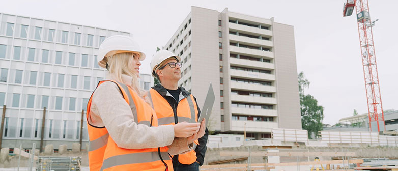 Eine Frau und ein Mann in Leuchtwesten stehen mit einem Notebook auf einer Baustelle