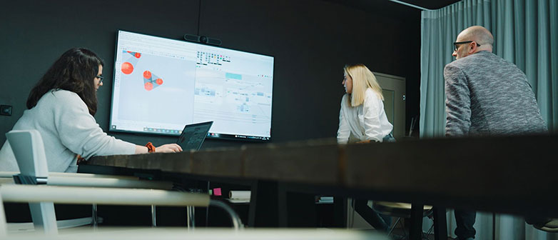 Zwei Frauen und ein Mann schauen in einem Büroraum auf einen grossen Bildschirm 