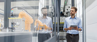 Mann in hellblauem Hemd hält Tablet in der Hand und begutachtet durch eine Glasfront mehrere gelbe Roboter