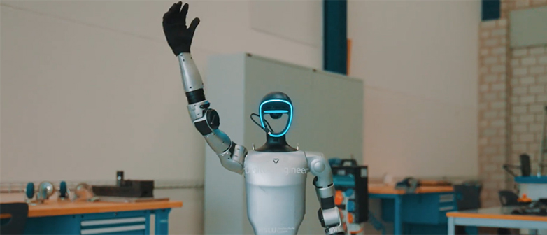 Humanoider Roboter mit leuchtendem Gesicht hebt eine Hand in einer technischen Werkstatt.