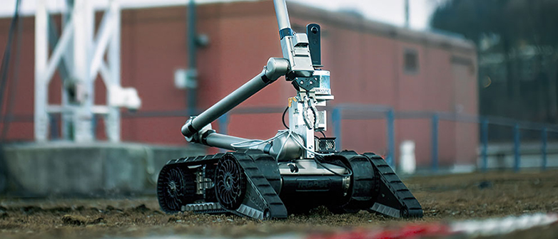 Ein Roboter mit Raupen steht auf einem bemoosten Hausdach, im Hintergrund ist ein rötliches Gebäude zu sehen