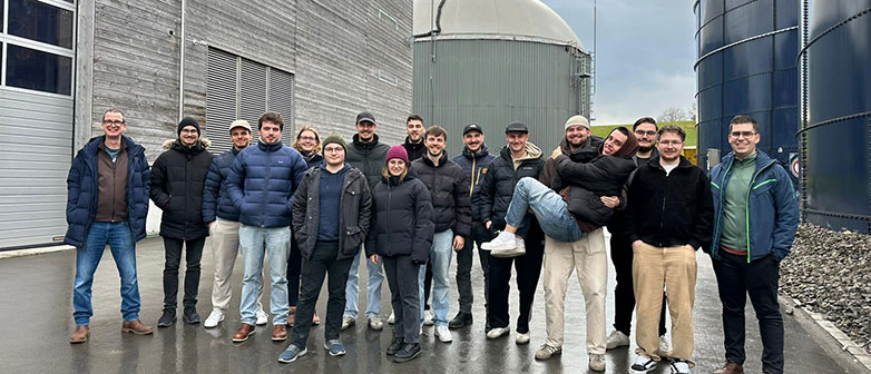 Studierendengruppe vor einer Biogasanlage mit Fermenter und Silotanks während einer Exkursion.