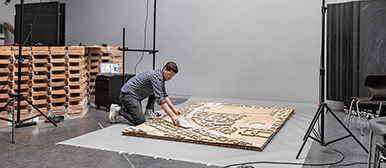 Ein Student kniet auf dem Boden eines Studios und arbeitet an einem grossen, flachen Modell aus Holz