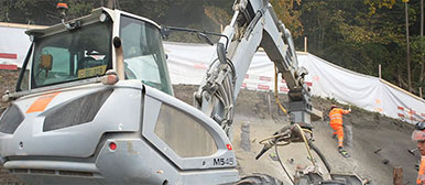 Bagger und Bauarbeiter in Warnkleidung bei Erdarbeiten auf einer Baustelle mit Böschung und Hangsicherung.