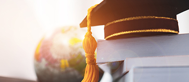 Ein Abschlusshut auf gestapelten  Büchern, im Hintergrund eine Weltkugel in hellem Sonnenlicht zu sehen