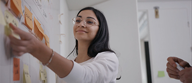 Eine braunhaarige Studentin mit weissem Pullover klebt lächelnd ein Post-it auf ein weisses Board