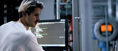 Ein Student mit weissem Shirt ist beschäftigt in einem technischen Raum mit Bildsdchirm und Technik im Hintergrund