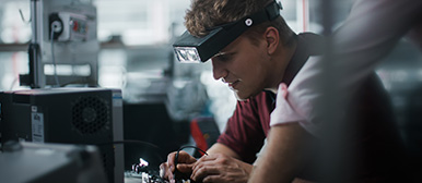 Ein Student, der eine Stirnlampe trägt, arbeitet konzentriert mit Elektrik, im Hintergrund sind weitere technische Gegenstände zu sehen